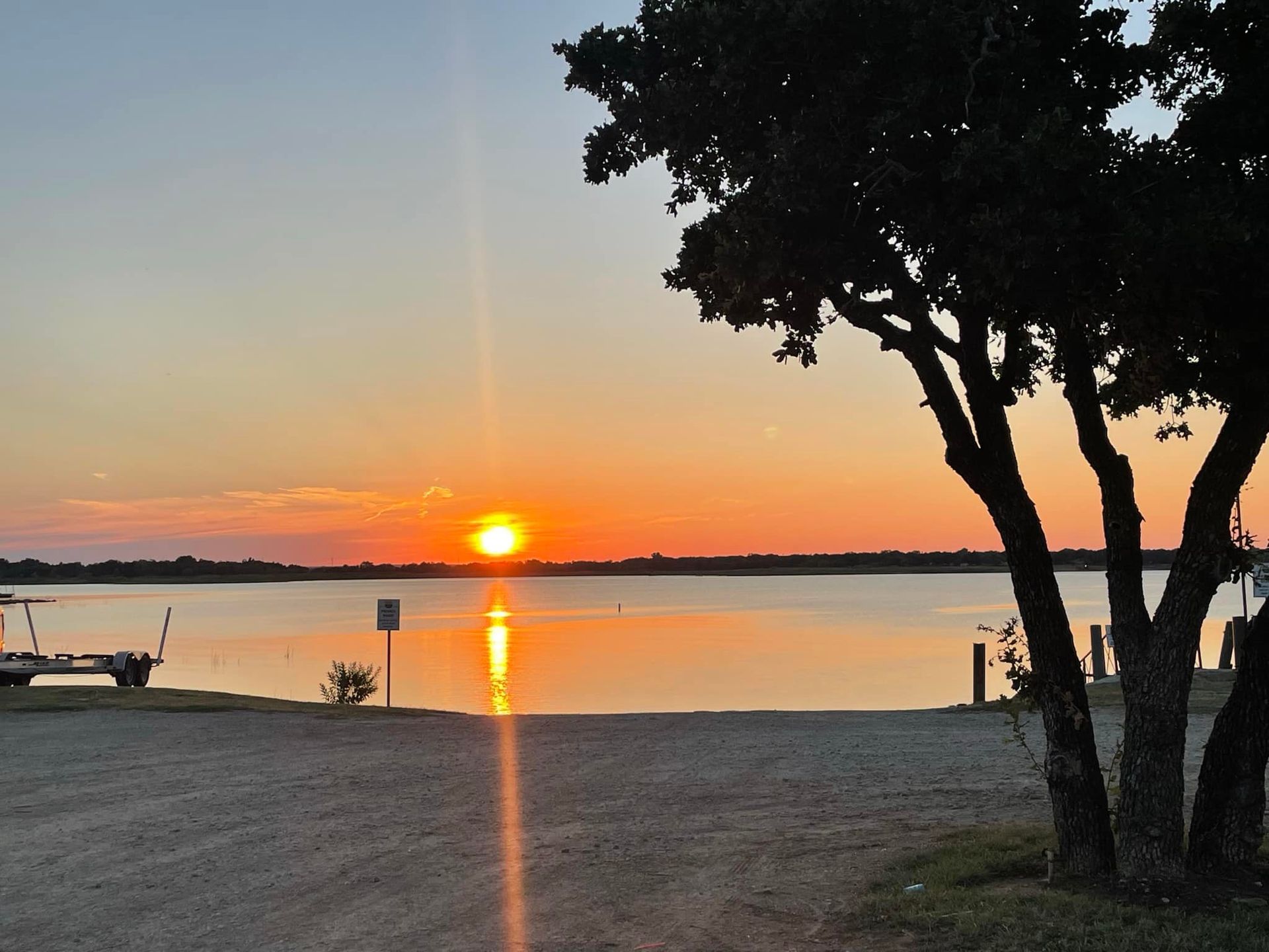 Sunset over a lake with a silhouetted tree in the foreground. Orange sun reflecting on the water and light blue sky.