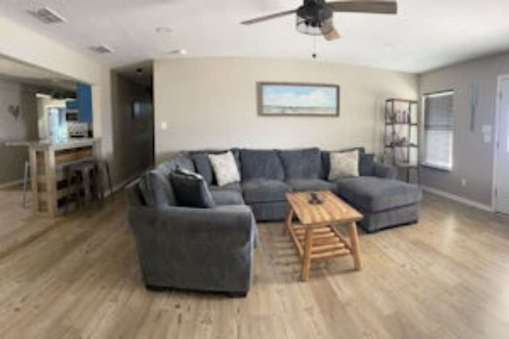 Spacious living room with a gray sectional sofa, wooden coffee table, and a mounted landscape painting. Wooden floors and a ceiling fan are visible.