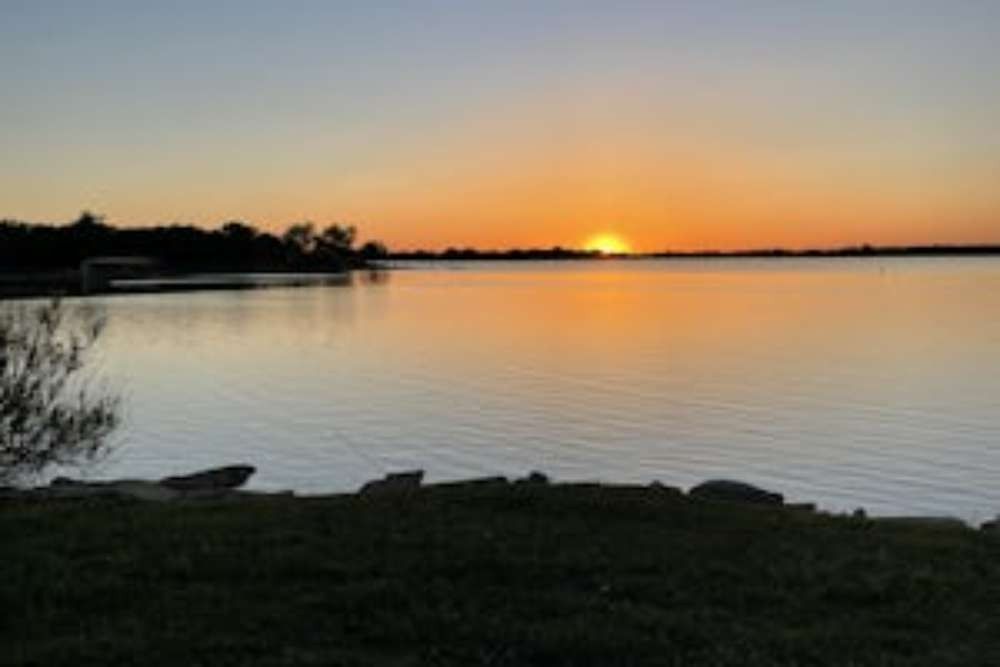 Sunset over a calm lake, reflecting orange and yellow hues. Silhouetted trees and shoreline in the background.