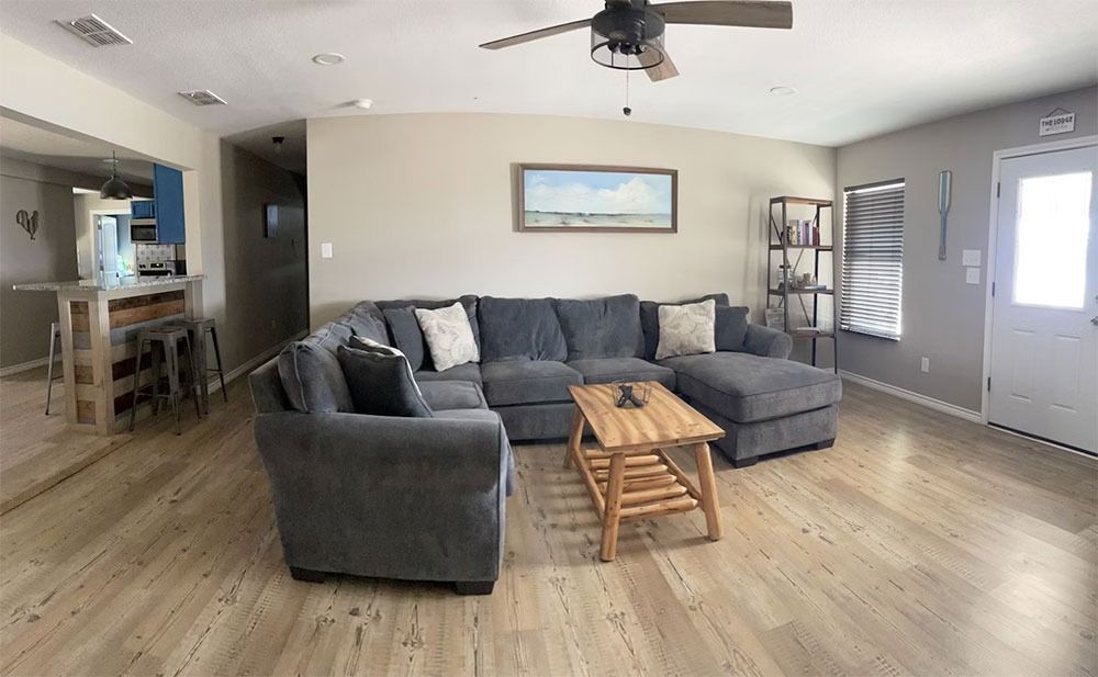 A gray sectional sofa and wooden coffee table in a living room with light wood floors and a blue kitchen in the background.
