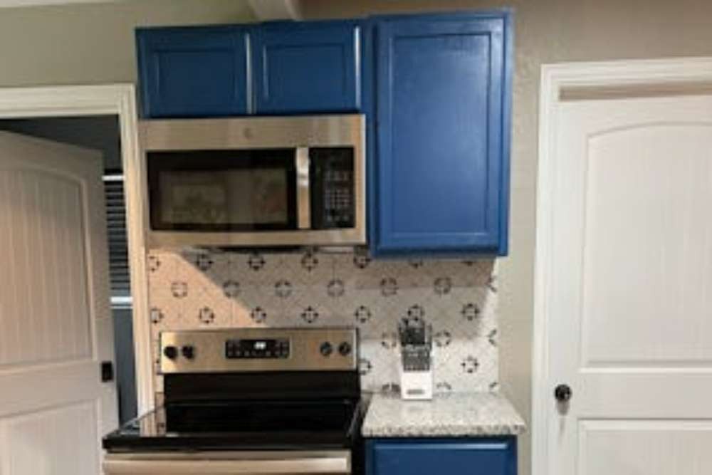 Blue kitchen cabinets with a stainless steel microwave and stove, white doors on either side, and a decorative backsplash.