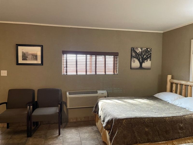 A motel room with a double bed, two chairs, a window with blinds, and wall art. Brown walls and a stone-tiled floor.