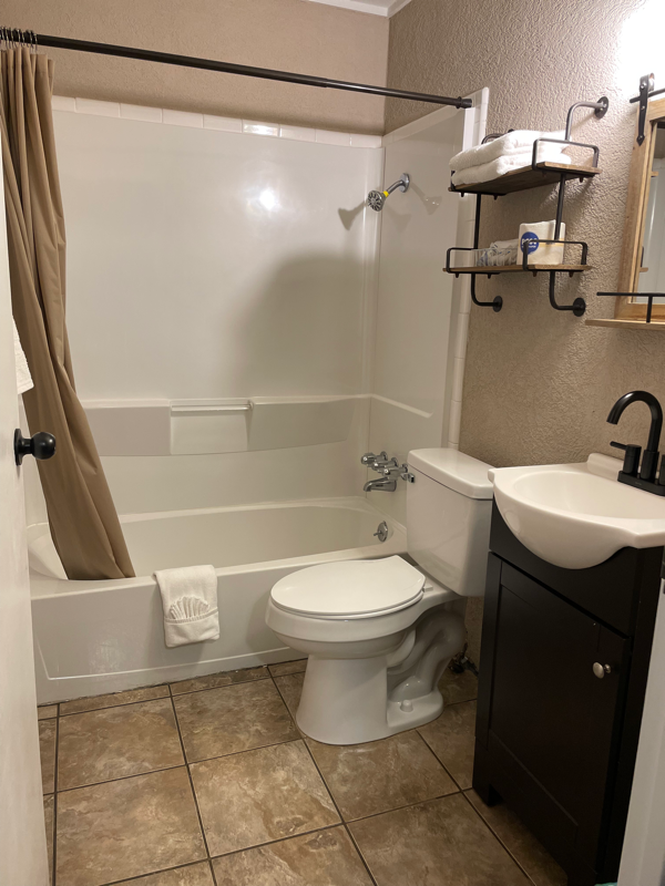 A small bathroom with a white tub and toilet, brown tiled floor, and dark vanity.