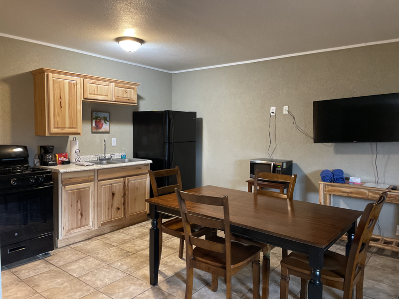 A small kitchen and dining area. Features wood cabinets, a black refrigerator and stove, a dark dining table with chairs, and a wall-mounted TV.