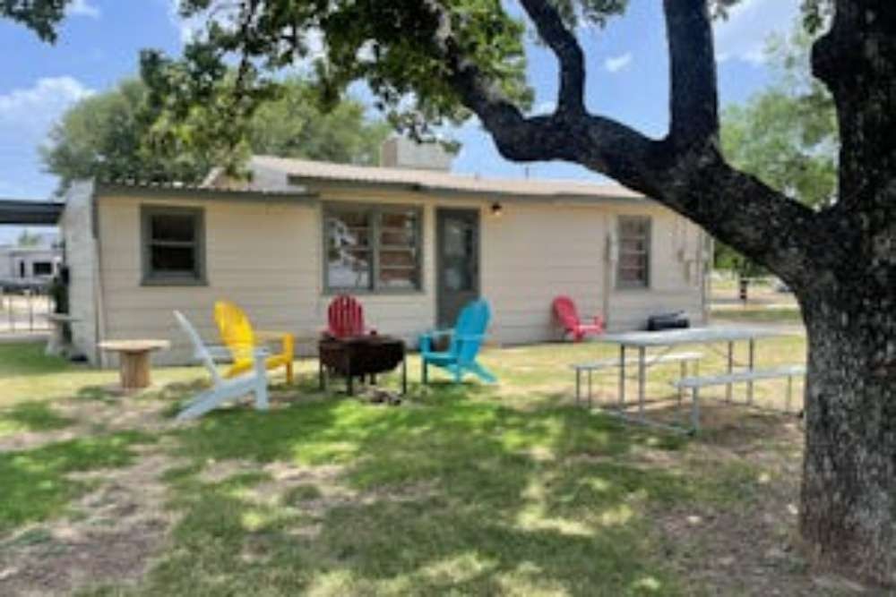 A small beige cabin with colorful chairs and a fire pit in a grassy yard, shaded by a tree, picnic table nearby.