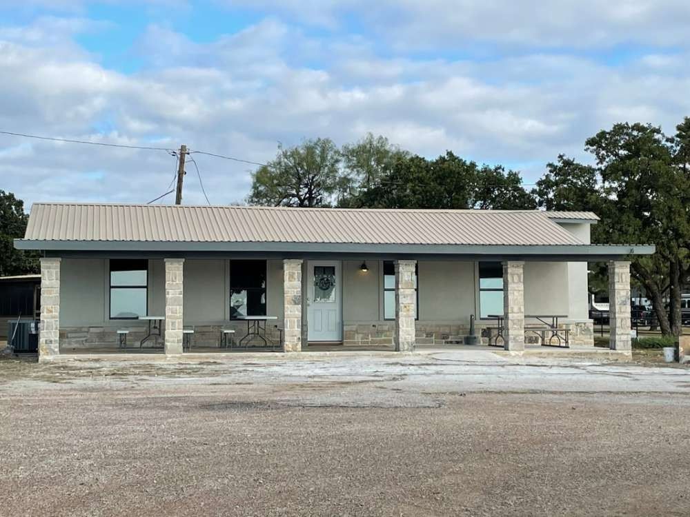 A single-story commercial building with a light-colored facade, columns, and a corrugated metal roof. The building sits on a gravel lot under an overcast sky.