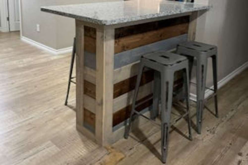 A rustic kitchen island with a granite countertop, wooden paneling, and two gray metal bar stools.