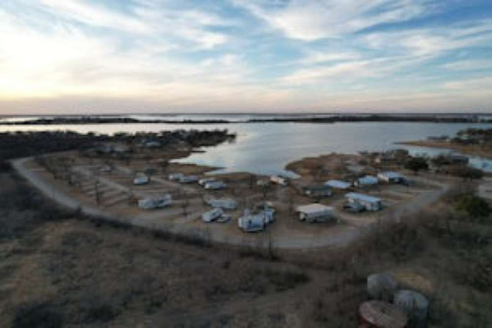 An aerial view of a lakefront RV park with RVs parked along a winding road. The sky is cloudy and the landscape is brown.
