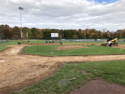 Baseball field under construction with dirt infield, grass outfield, and a small excavator on the right