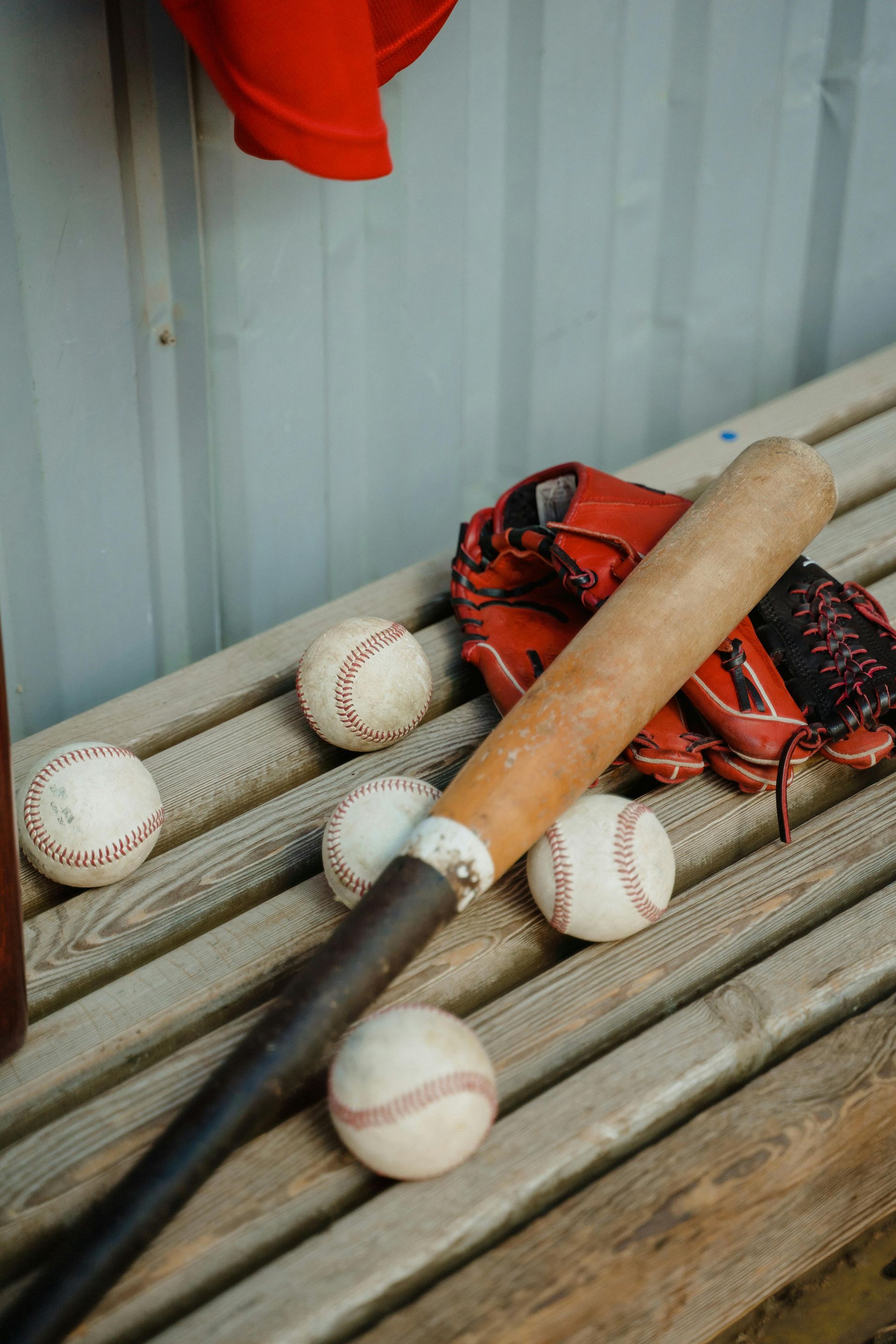 Baseball bat, glove, and several balls on a wooden bench against a corrugated wall.