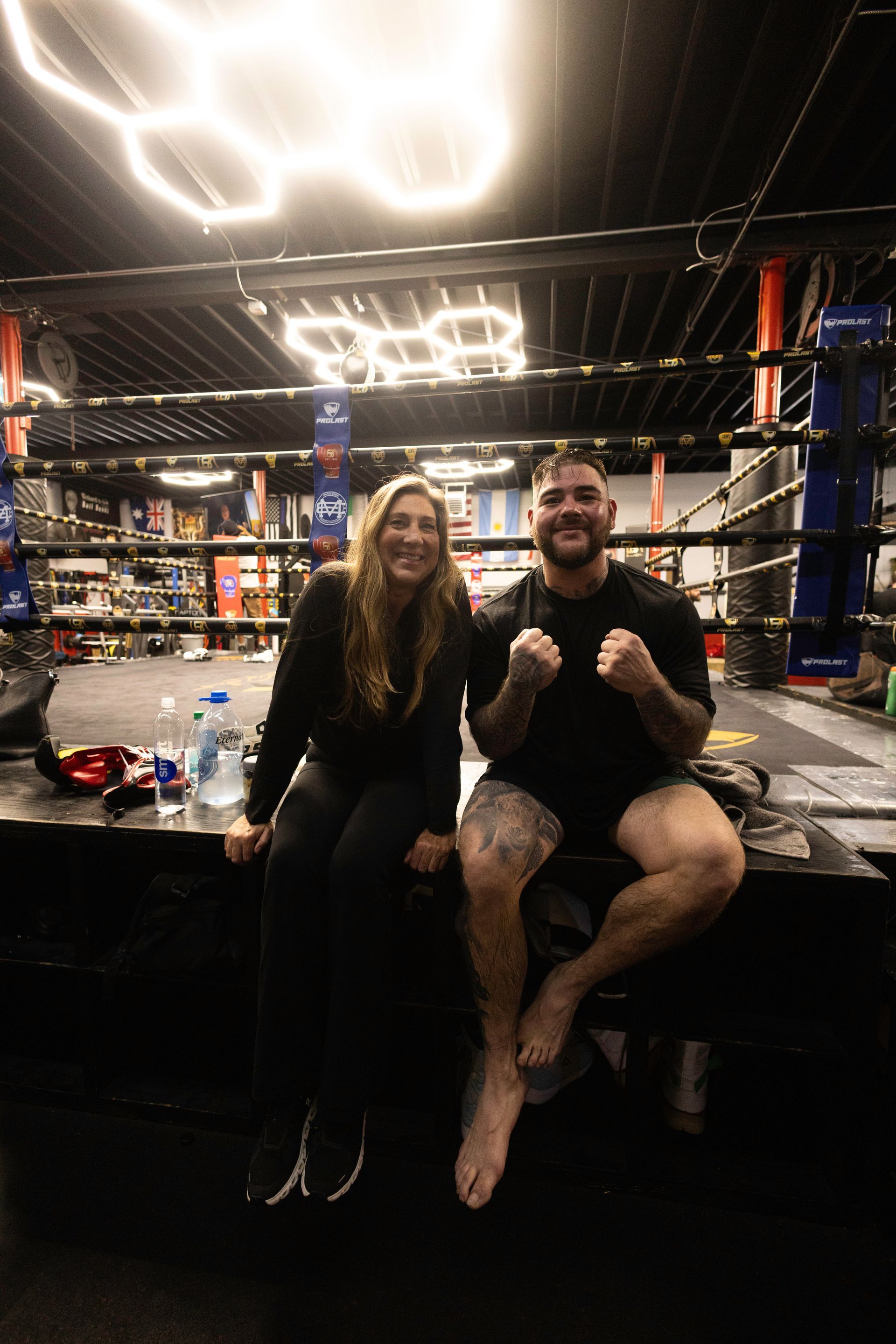 Andy Diaz and Evelyn Hallford sitting in a boxing ring, smiling with fists raised under bright gym lights