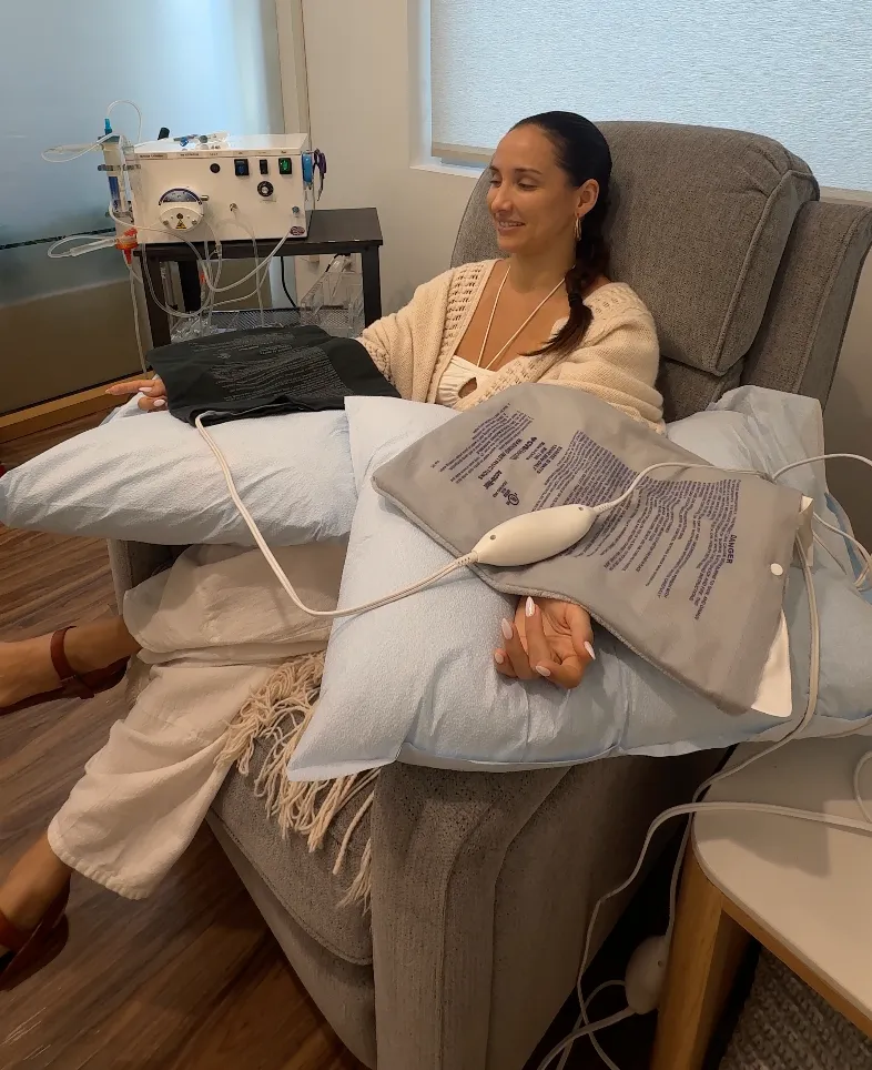 Woman receiving EBOO therapy while seated in a gray recliner with a towel and spa equipment nearby