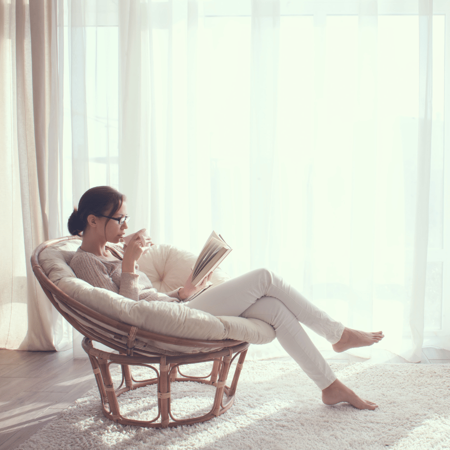 Woman reading in a wicker chair by a bright window, with soft white curtains and a cozy room.
