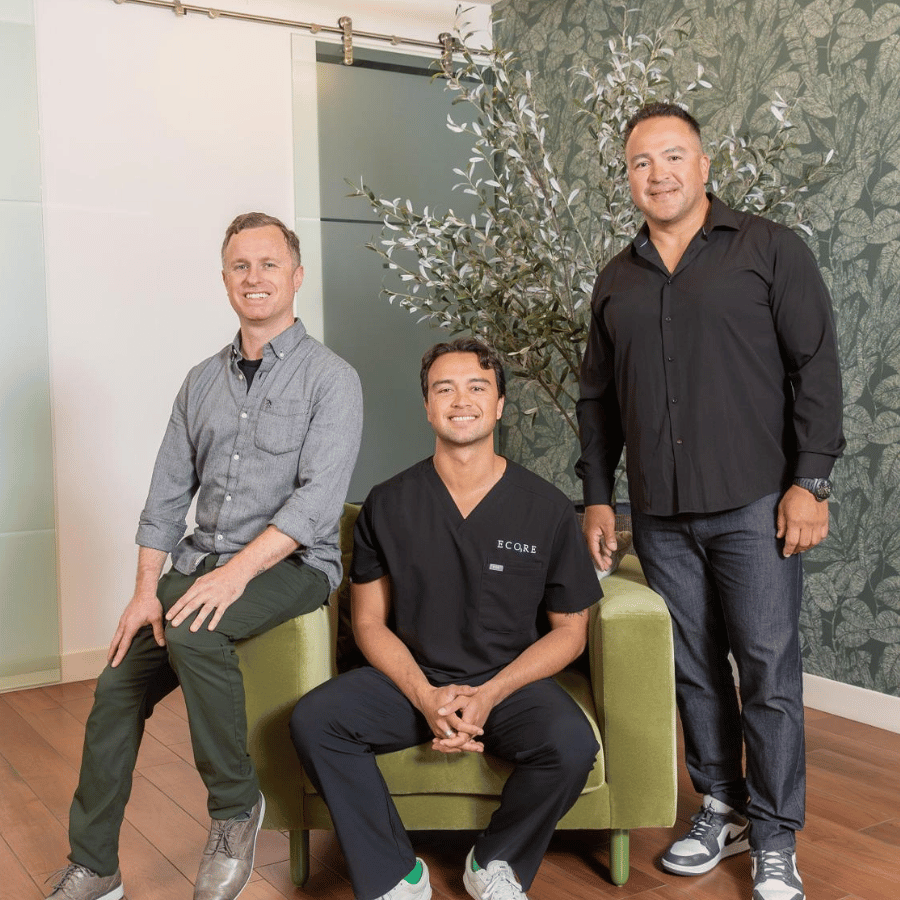 Three men seated in a modern office lounge, posing on green chairs beside a plant