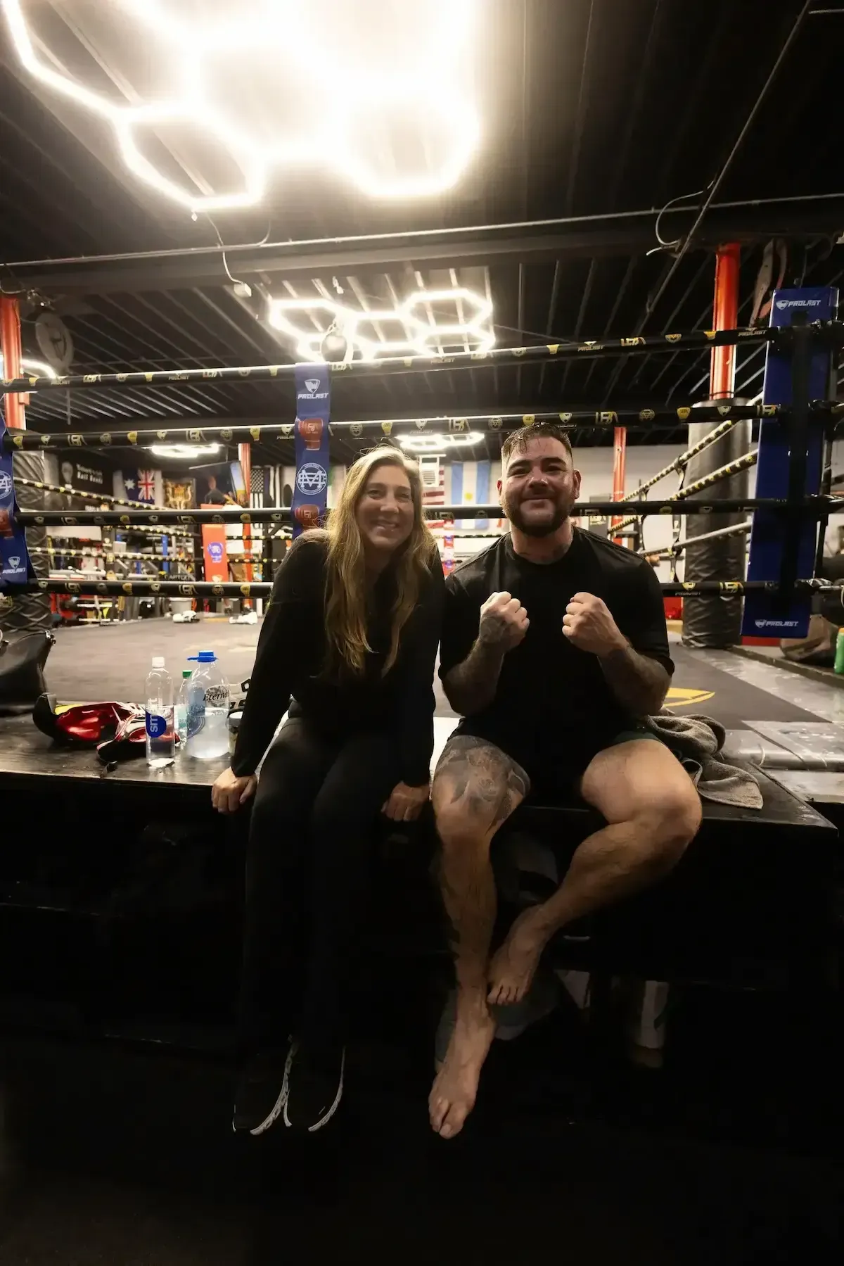 Andy Diaz and Evelyn Hallford sitting in a boxing ring, smiling with fists raised under bright gym lights