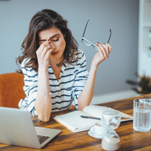 Woman at a desk rubbing her eyes while holding glasses, with a laptop, notebook, and coffee cup nearby