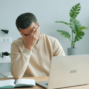 Person at desk covering face beside an open laptop and notebook, looking stressed