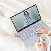 Video call with a doctor on a laptop while someone holds a mug on a white bed