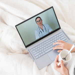 Video call with a doctor on a laptop while someone holds a mug on a white bed