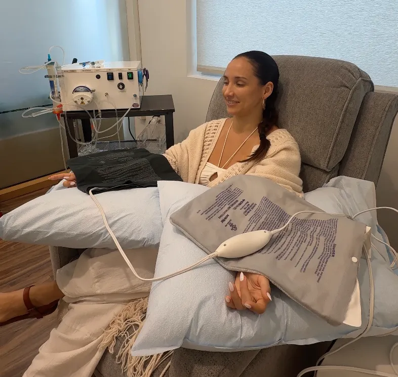 Woman receiving EBOO therapy while seated in a gray recliner with a towel and spa equipment nearby