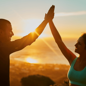 Two people high-five at sunset by the ocean, silhouetted against a golden sky.