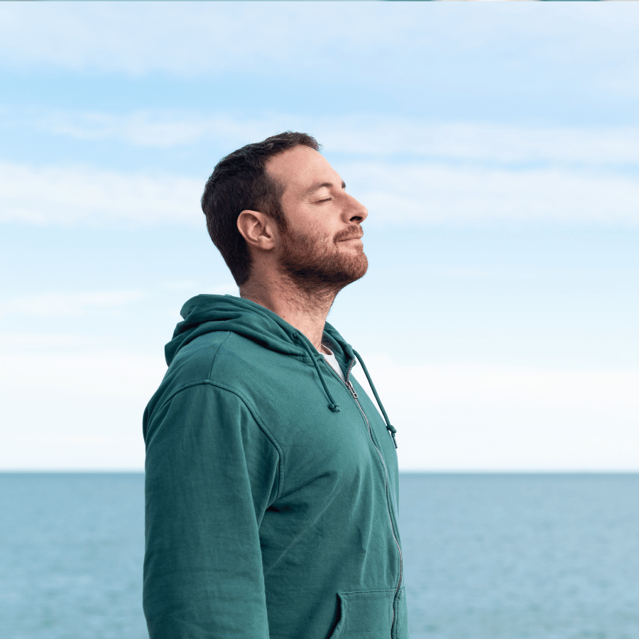 Man in a green hoodie standing by the sea, eyes closed, with a calm expression under a blue sky