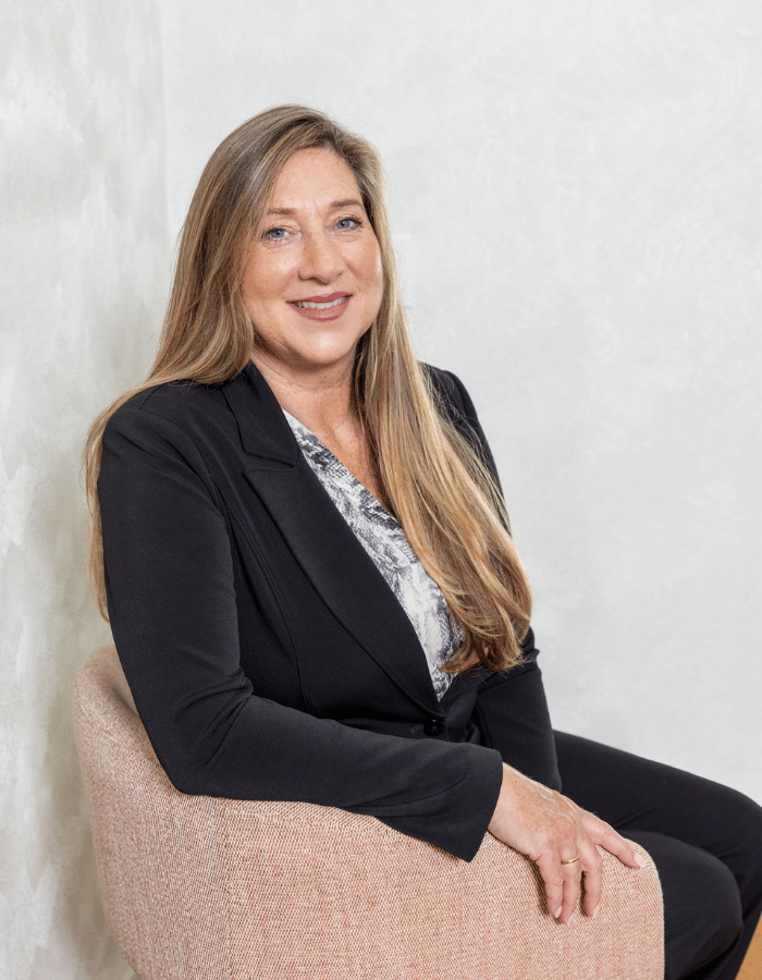 Woman seated in a blush chair, smiling in a black blazer against a light gray background.