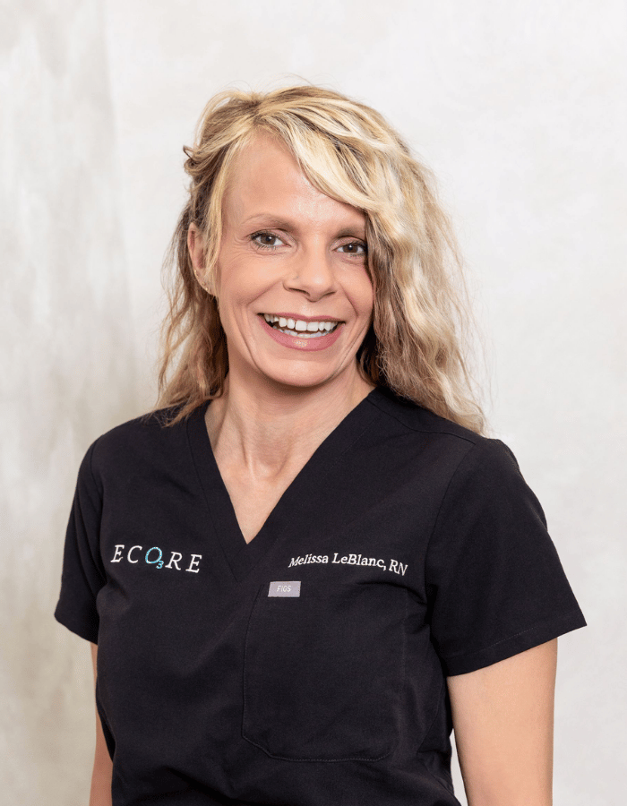 Smiling person in black scrubs with logos, standing against a light background.