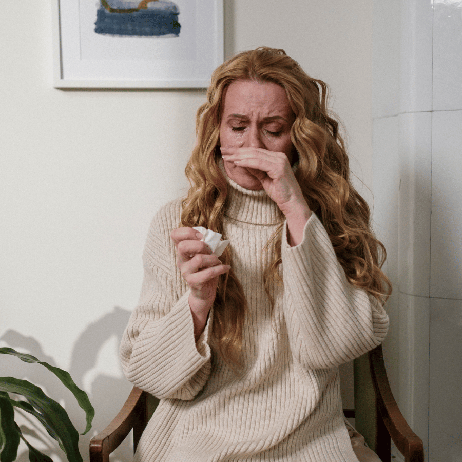 Woman in a beige sweater coughing into her hand while holding a tissue indoors.