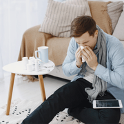 Person sitting on a couch, blowing their nose beside medicine and a cup in a cozy living room