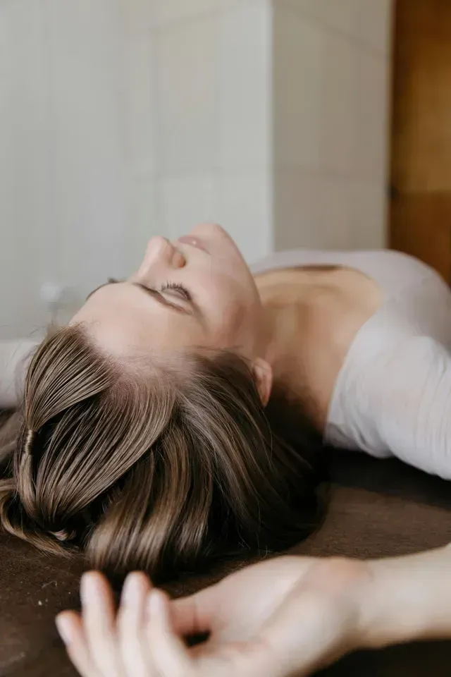 Woman reclining on a wooden floor with eyes closed, wearing a white top, hand in the foreground