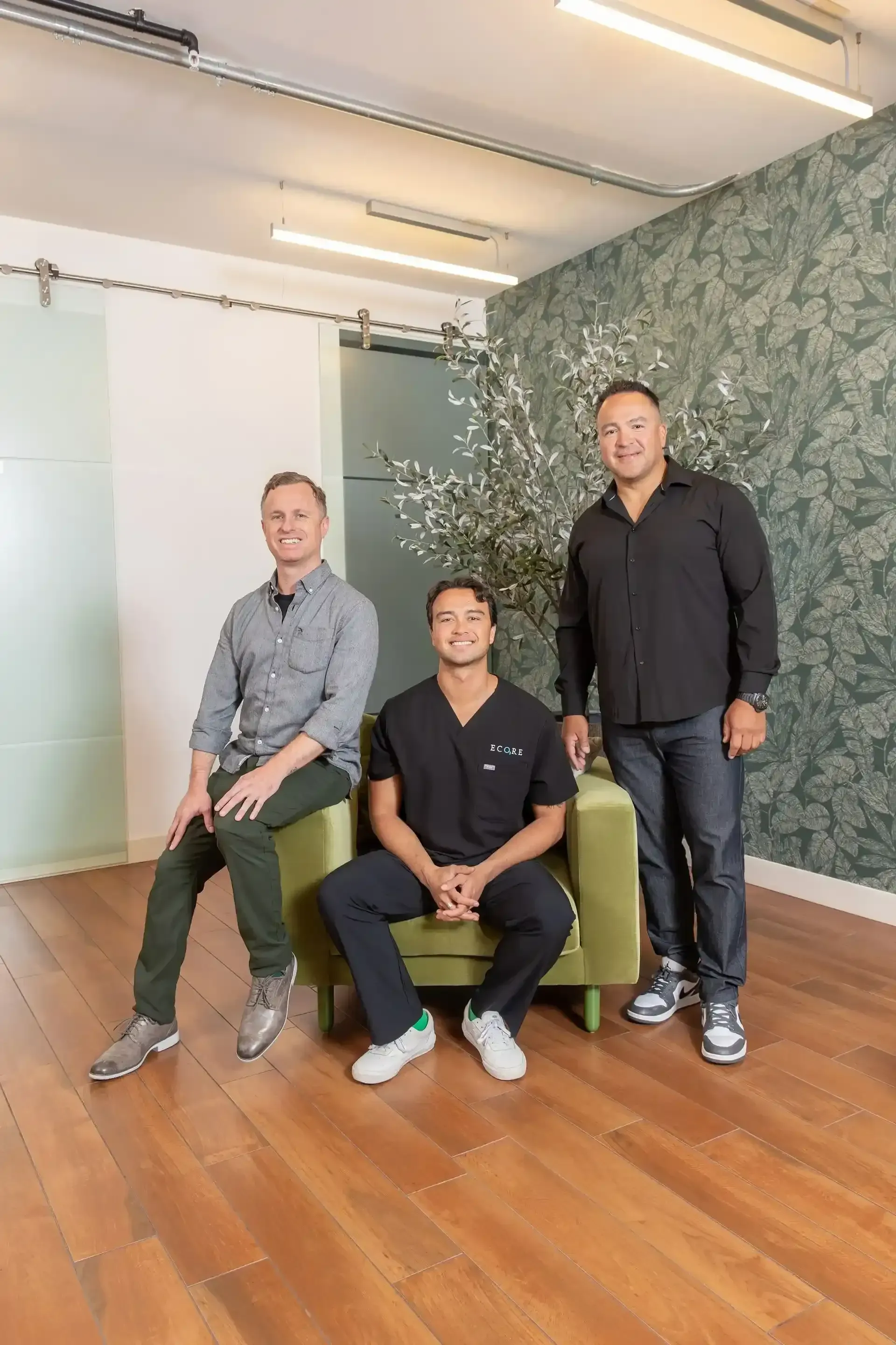 Three people posing in a modern office with wood floors, a plant wall, and bright overhead lights.