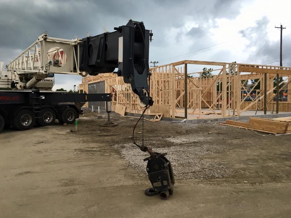 Construction crane lifting a beam on a muddy site beside wooden house framing.