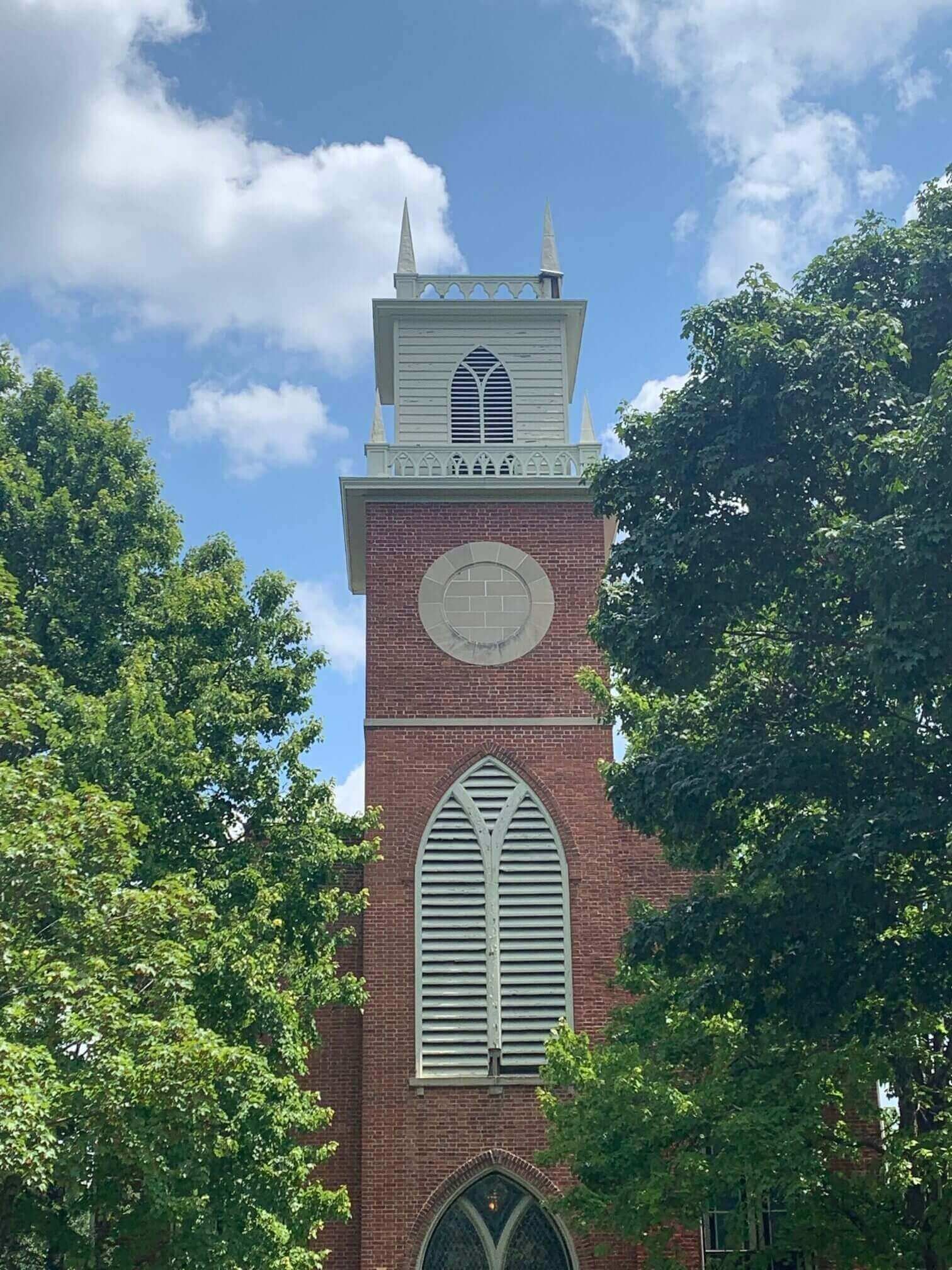 Red brick church tower with white steeple framed by green trees and blue sky.