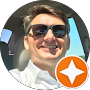 Homem de óculos escuros sorrindo, dirigindo um carro com um logotipo de estrela laranja.