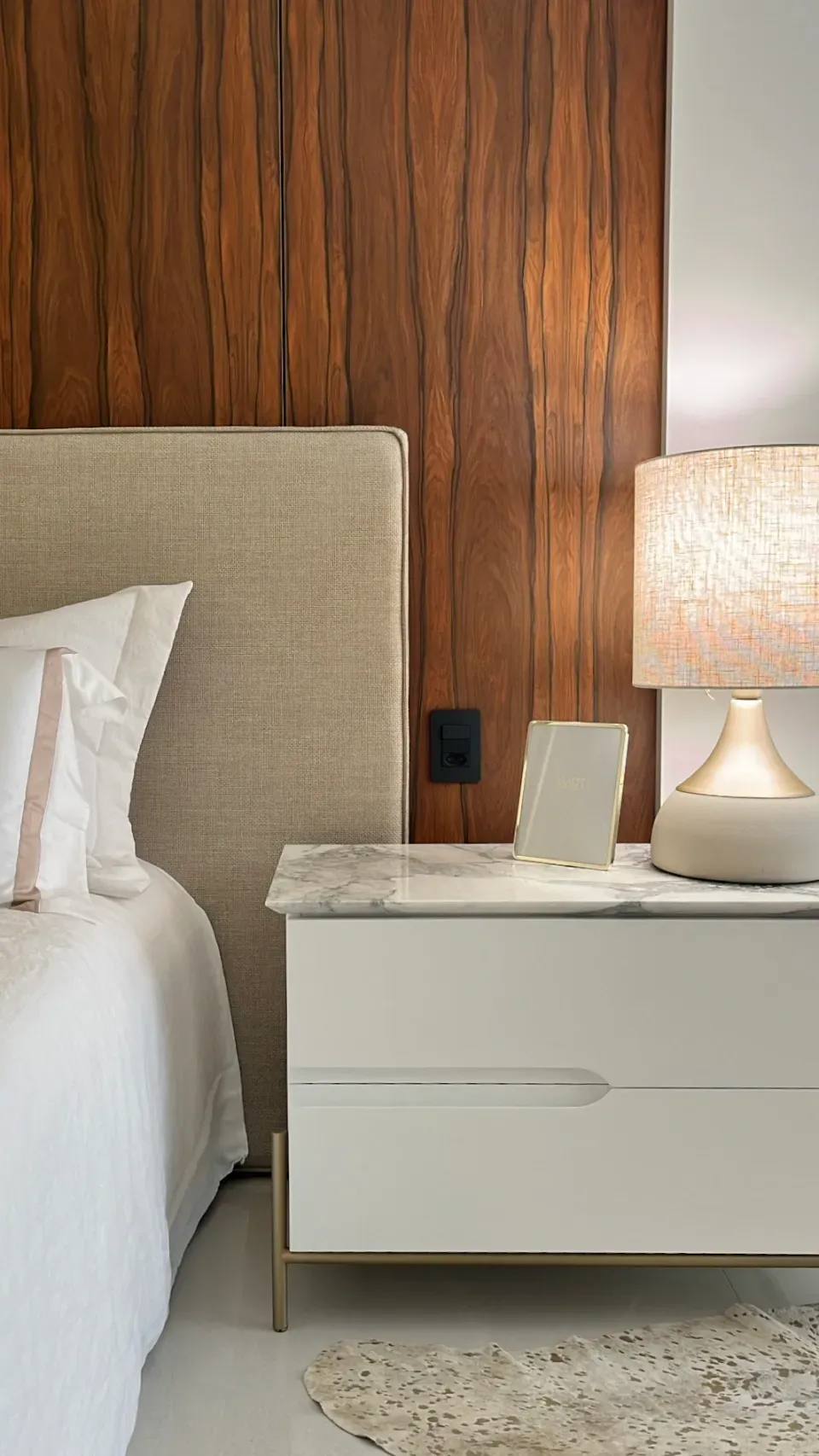 Quarto com uma parede de destaque em madeira, uma cama com cabeceira bege e uma mesa de cabeceira branca com um abajur.