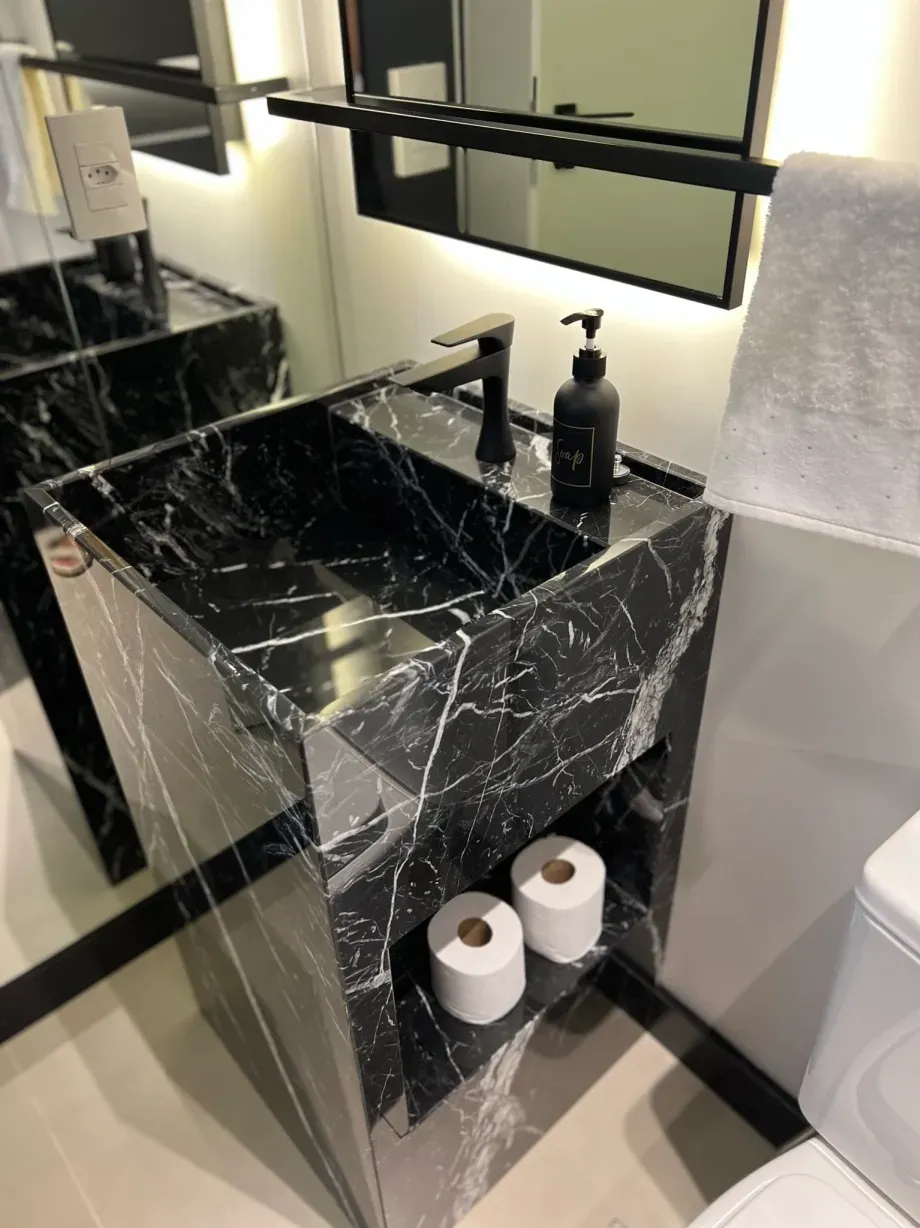 Bancada de banheiro em mármore preto com pia embutida e prateleira aberta para rolos de papel higiênico.