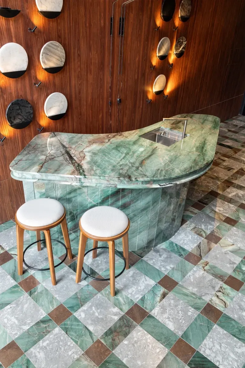 Bar com bancada de mármore verde e dois bancos, em contraste com madeira e azulejos estampados.