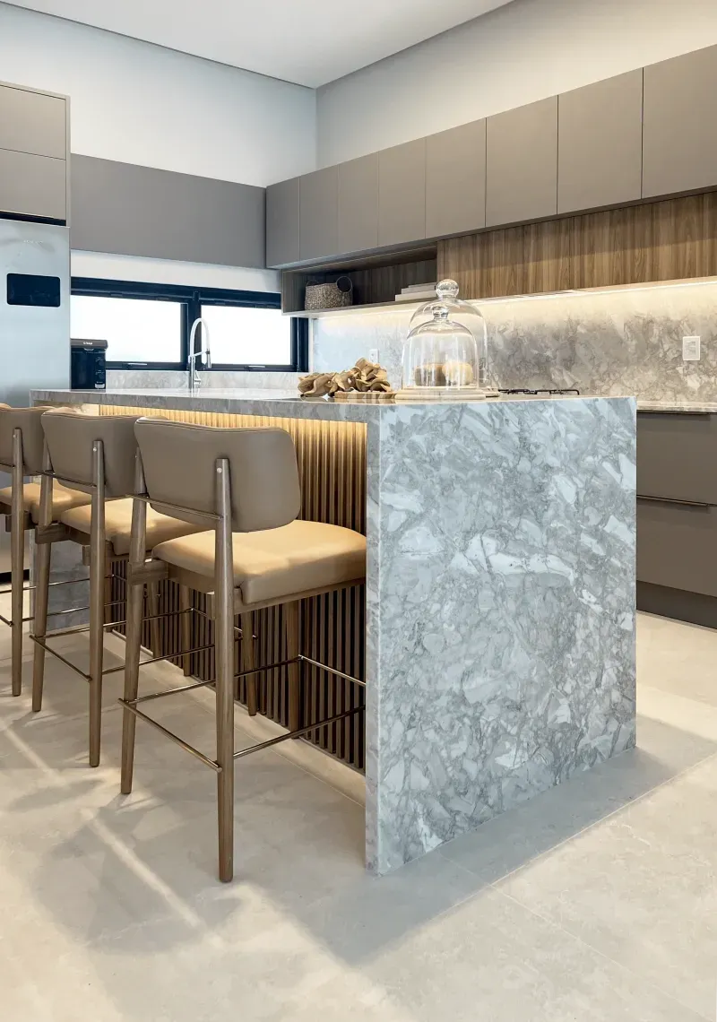 Cozinha moderna com ilha central com tampo de mármore e banquetas de bar em tom bege claro.