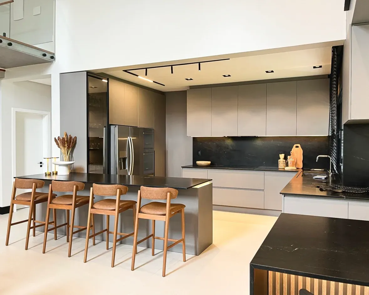 Cozinha moderna com ilha e banquetas, bancadas escuras, armários cinza e eletrodomésticos embutidos.