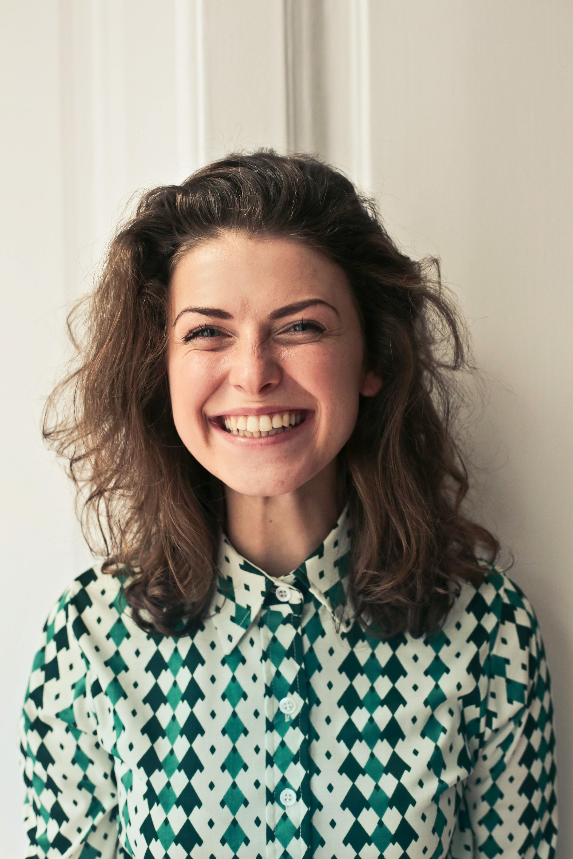 Mulher sorridente, vestindo uma blusa verde estampada, em pé em frente a uma porta branca.