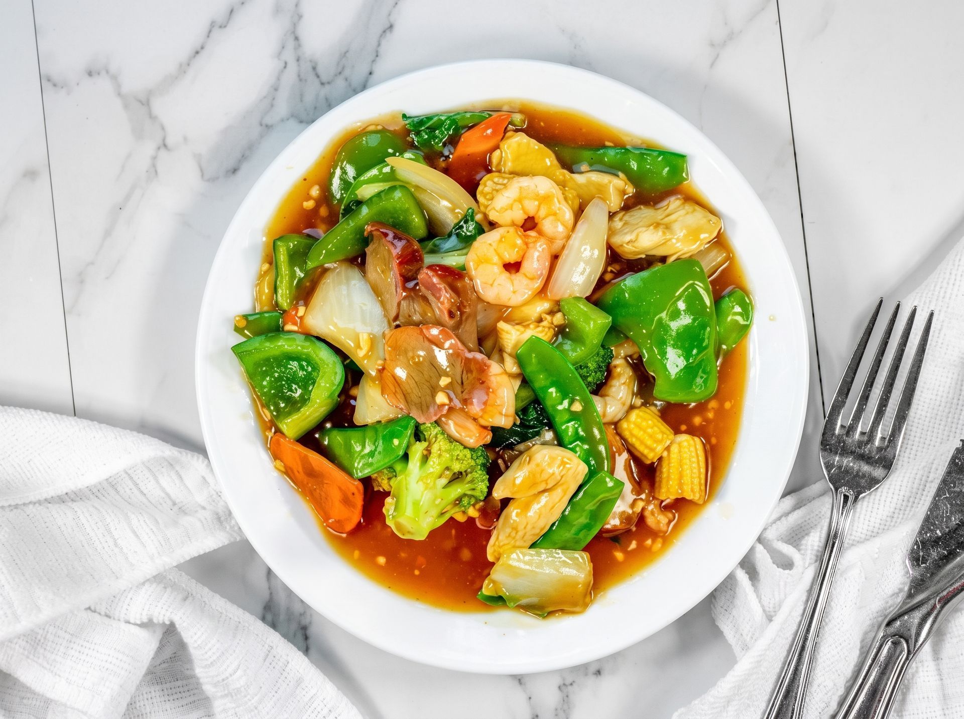 Stir-fried shrimp and vegetables in a white bowl on a marble table, with a fork and napkin nearby