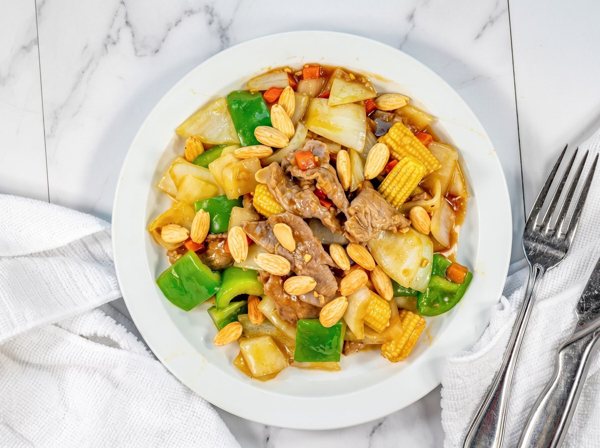 Bowl of mixed vegetable stir-fry with peanuts on a white table, beside a fork and napkin.