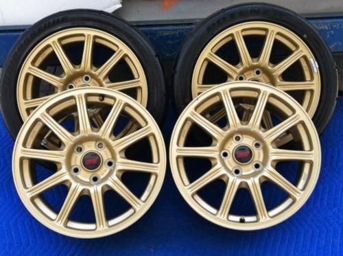 Four gold alloy car wheels with black tires on a blue mat, two stacked behind two upright wheels