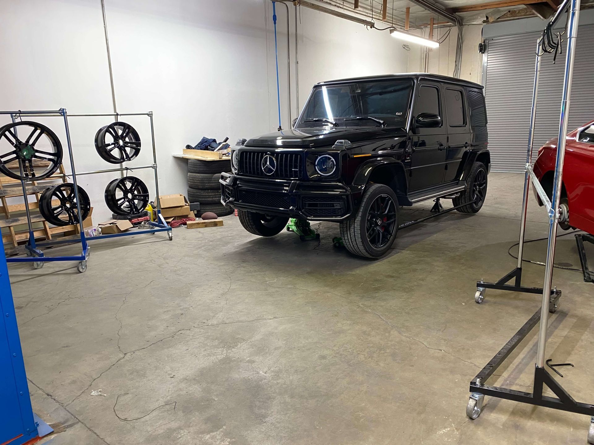 Black Mercedes G-Class SUV in a garage beside wheel racks and a red car.