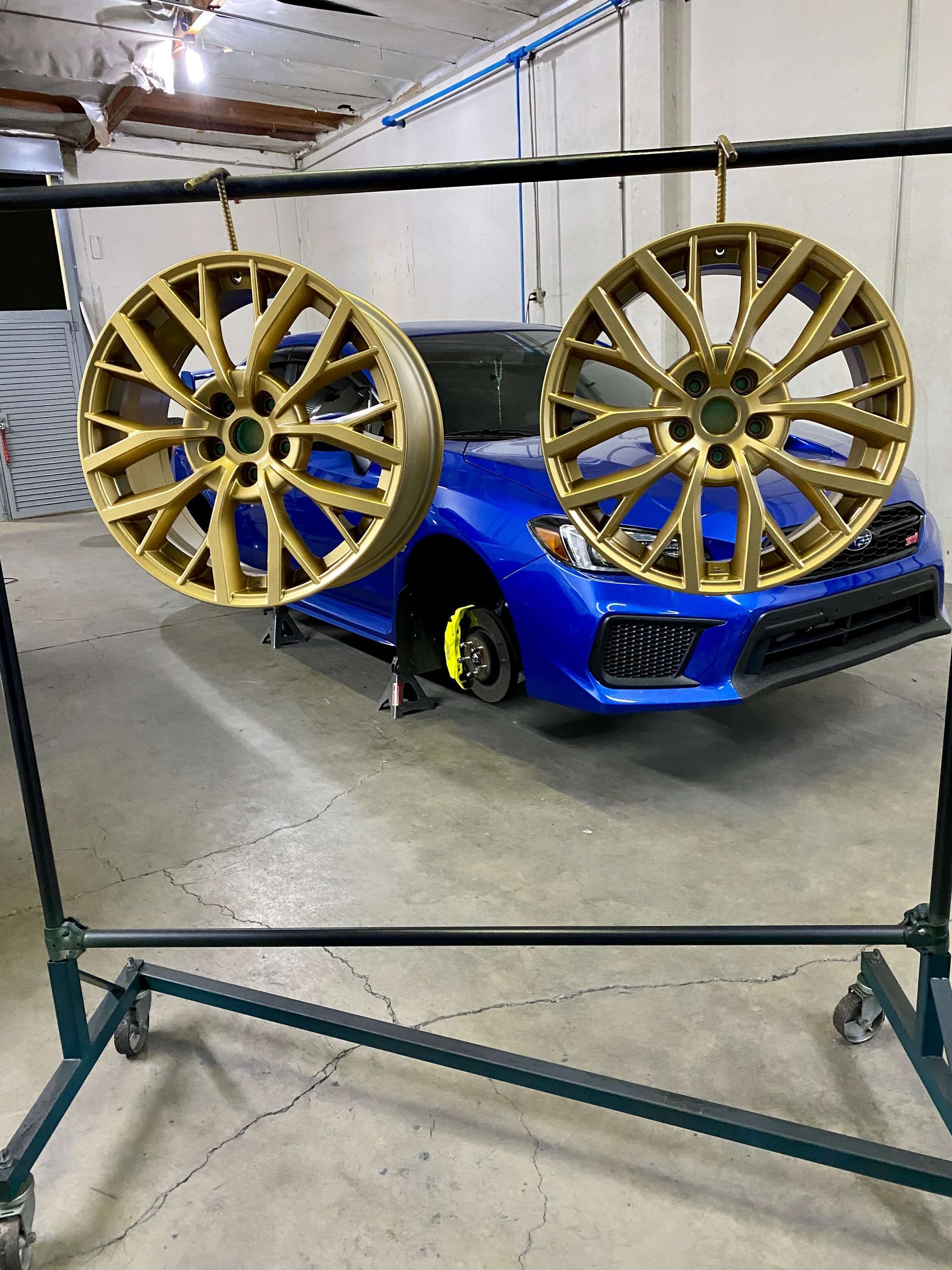 Blue sports car behind two gold alloy wheels on a display stand in a garage-like setting