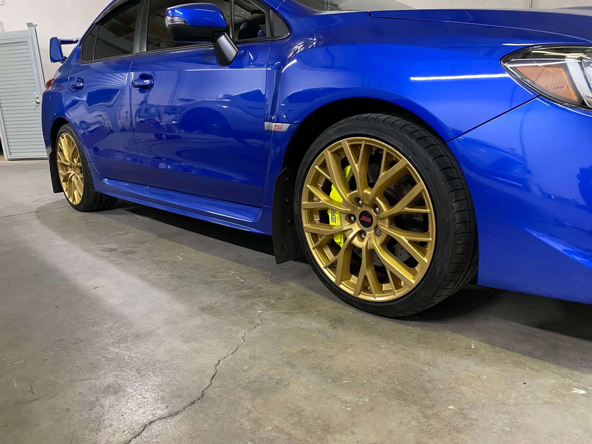 Blue sports car with gold rims parked indoors on a concrete floor.
