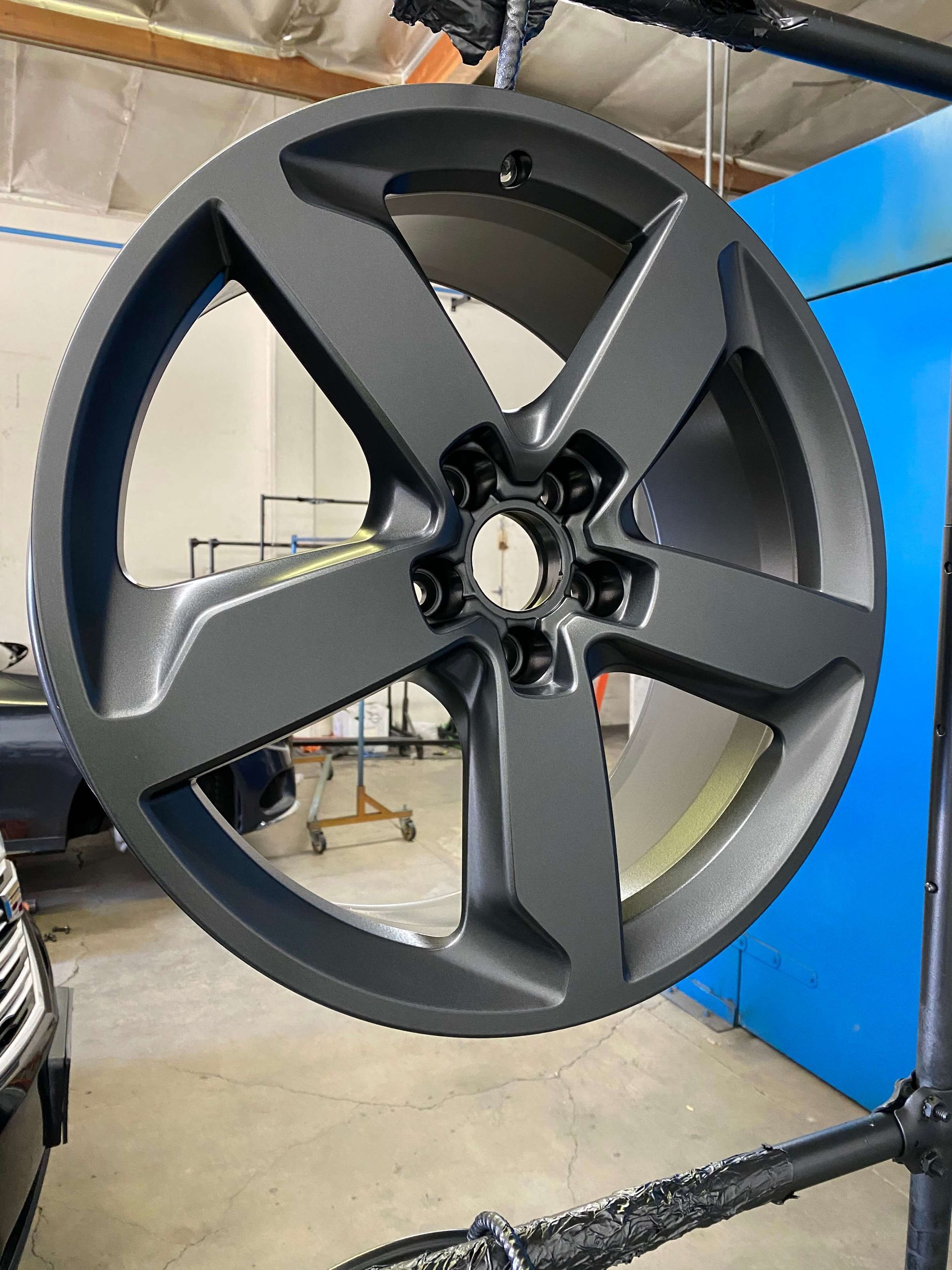 Matte black alloy wheel rim hanging in a workshop booth, with blue walls and a spray frame visible behind it.