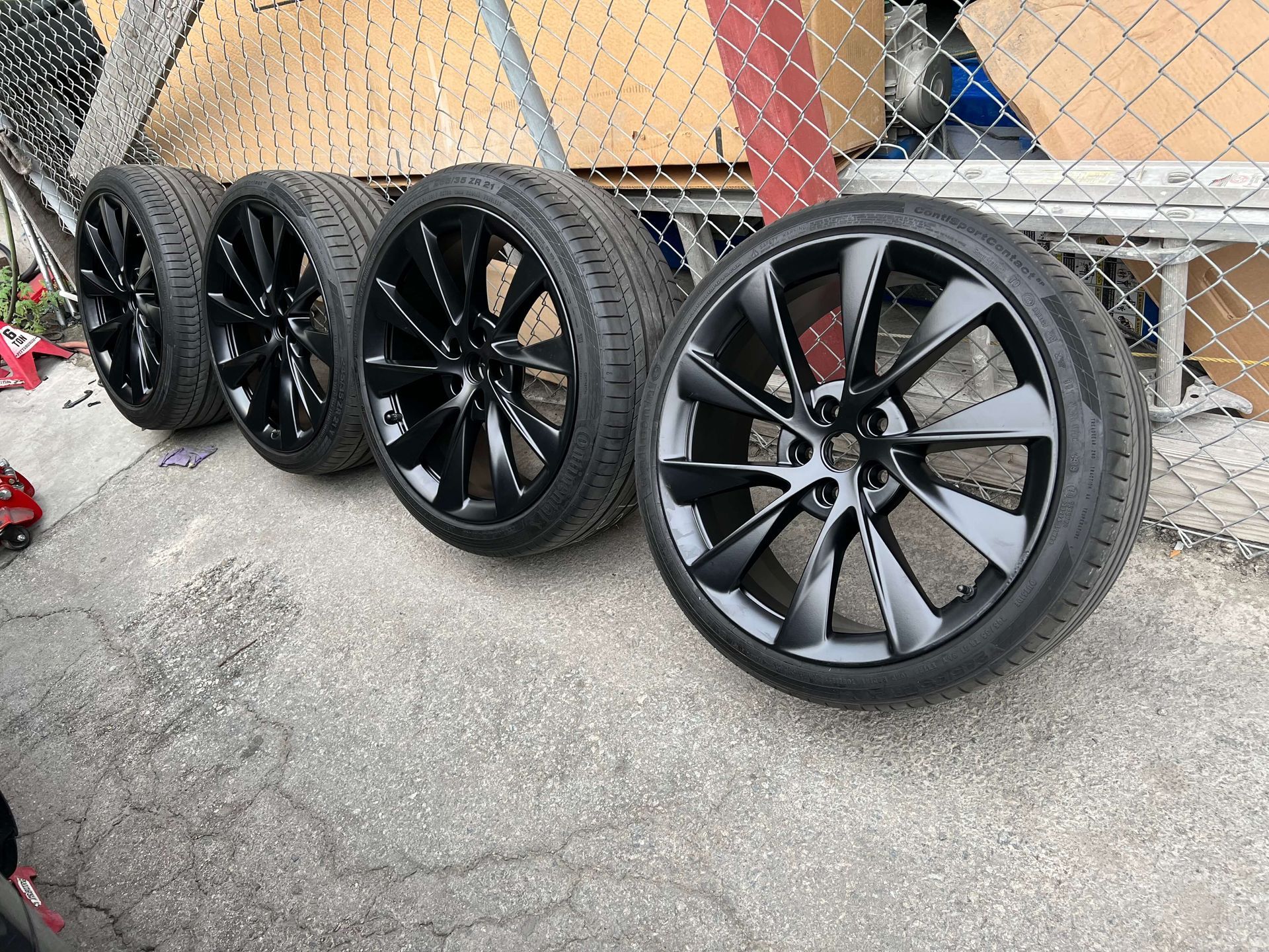Four black alloy car wheels with tires lined up on a concrete surface by a chain-link fence.