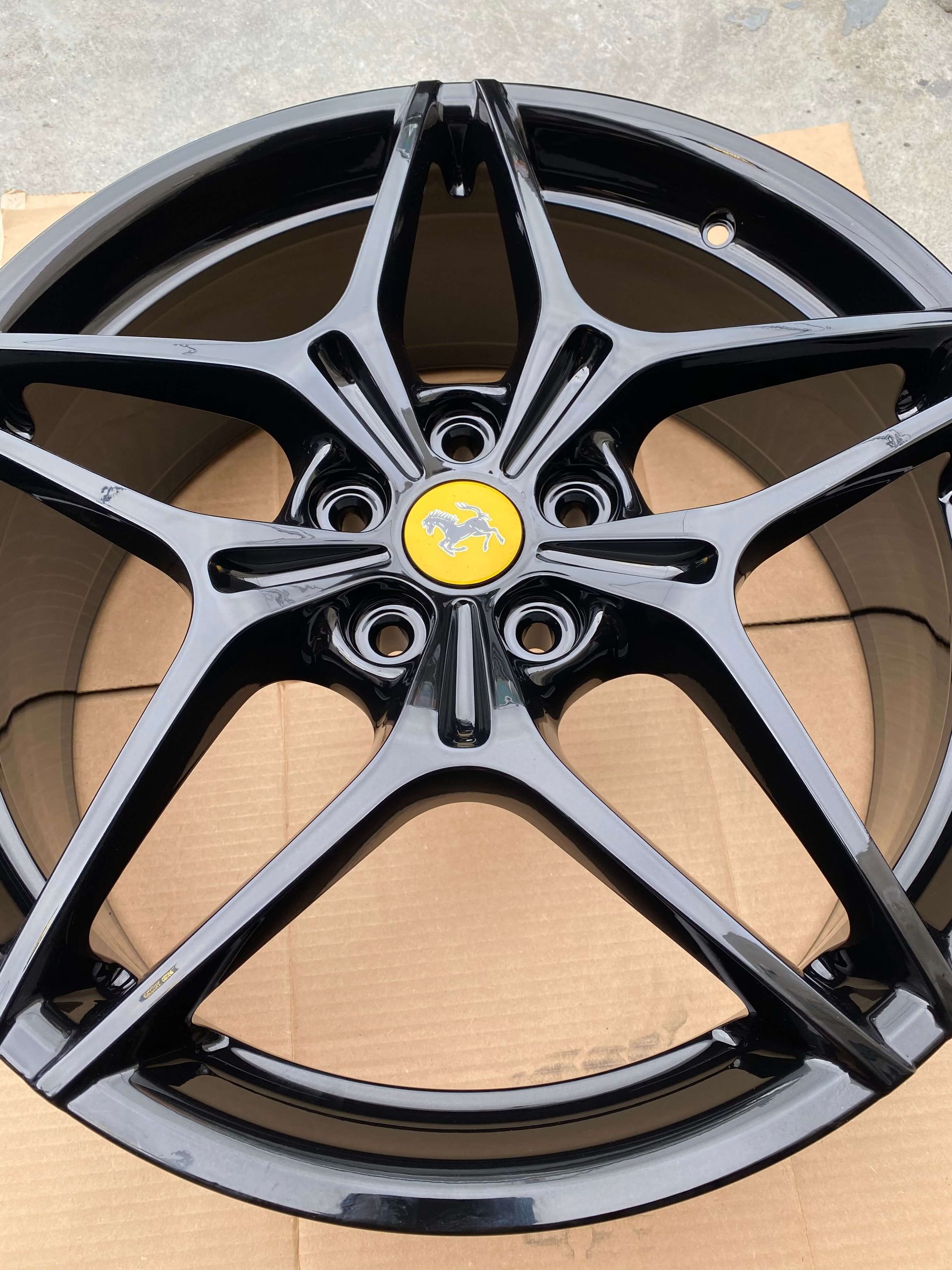 Black alloy car wheel rim with a yellow Ferrari center cap, shown on cardboard background
